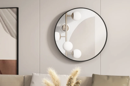 Round wall mirror with black frame reflecting light above beige sofa with cushions and pampas decor.