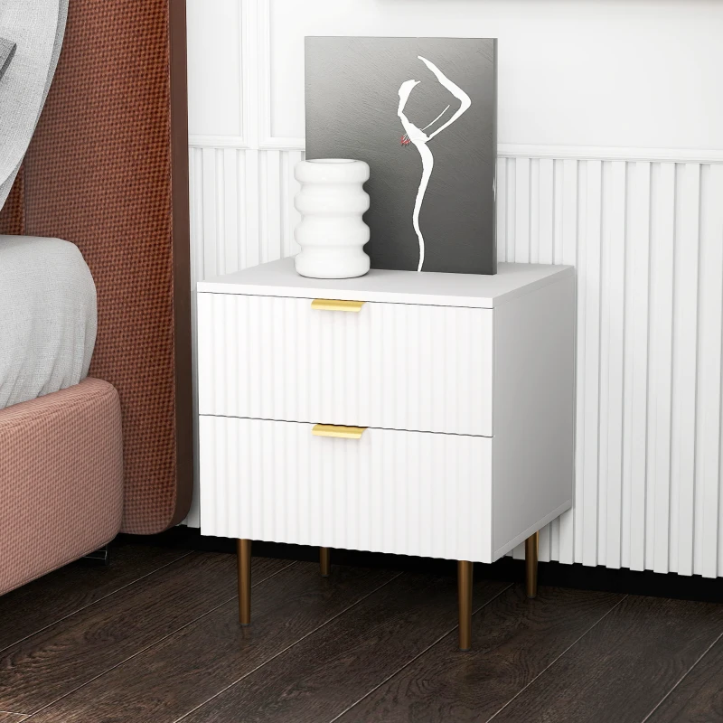 Modern white bedside table with two drawers, gold handles, metal legs, and decorative artwork.