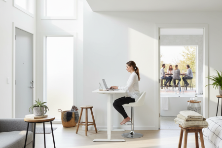 Woman working on laptop at tall white desk in bright home office.