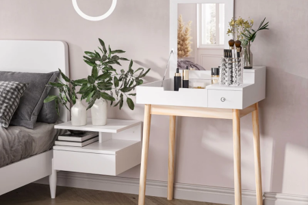 White dressing table with mirror, makeup items, and stool placed beside a bed in bright room.