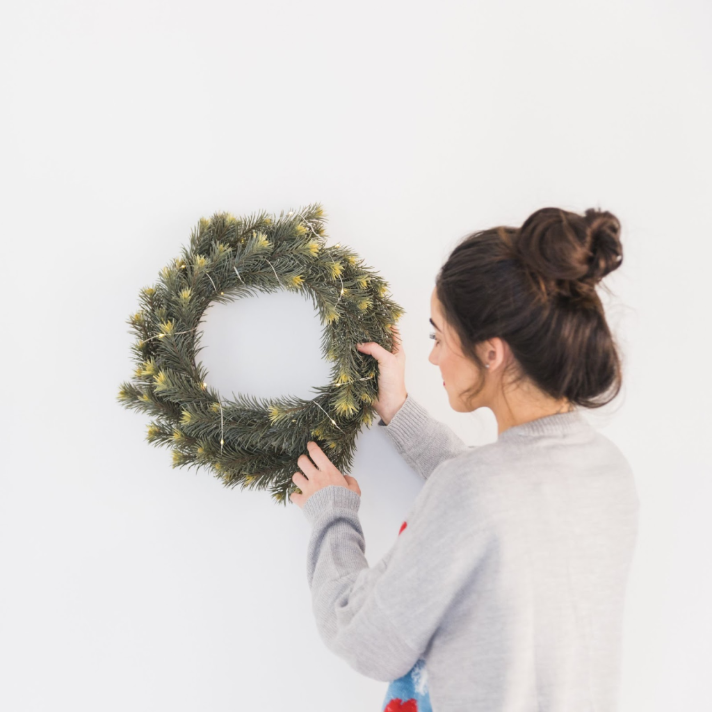 Woman hanging a lit Christmas wreath on a plain white wall indoors for festive decoration.