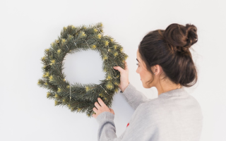 Woman hanging a lit Christmas wreath on a plain white wall indoors for festive decoration.