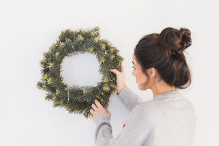 Woman hanging a lit Christmas wreath on a plain white wall indoors for festive decoration.