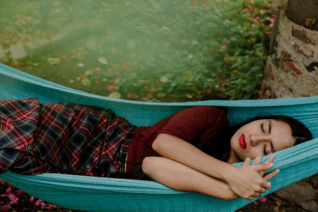 Woman sleeping on blue hammock outdoors surrounded by greenery.