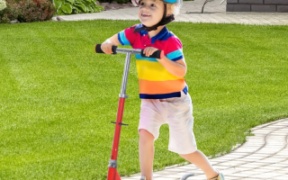 Young boy standing proudly with light blue scooter near lakeside homes.