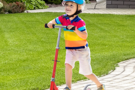 Young boy standing proudly with light blue scooter near lakeside homes.