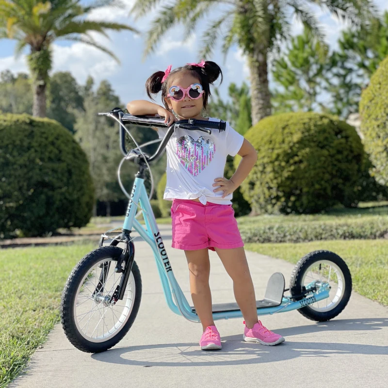 Little girl posing beside blue kick scooter on sunny walkway.