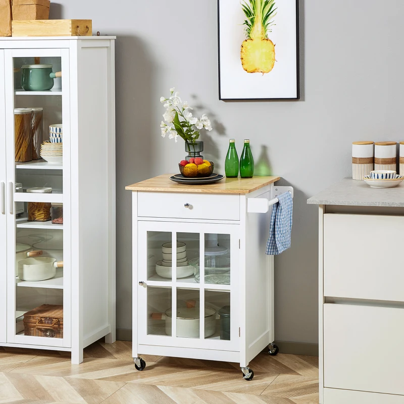  White kitchen cart with wooden top, fruit bowl, and flower vase beside glass cabinet and wall art.
