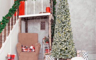 Snow-flocked slim Christmas tree beside gifts and cozy chair.