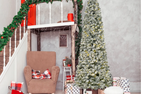 Snow-flocked slim Christmas tree beside gifts and cozy chair.
