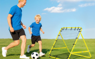 Father and son playing soccer with rebounder net on grassy field.