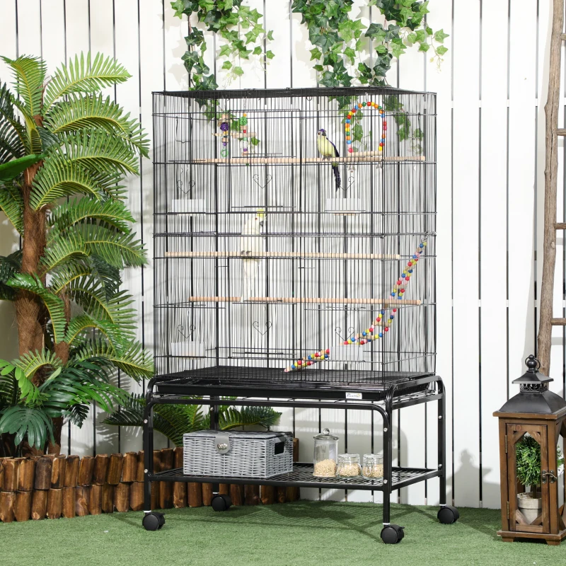 Large outdoor bird cage with colorful hanging toys and perches among tropical plants.