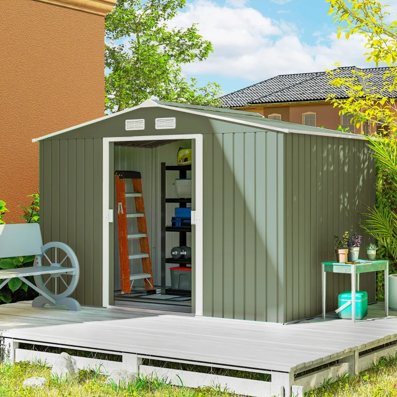 Light green metal garden shed near wooden deck surrounded by plants and a lawn mower inside.