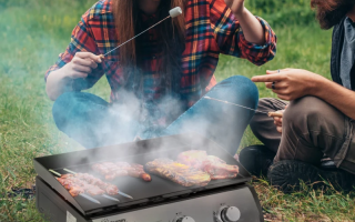 Couple grilling meat and marshmallows on portable Outsunny BBQ grill in grassy outdoor setting.