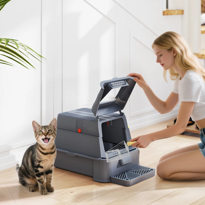 Woman cleaning grey litter box as tabby cat sits nearby.