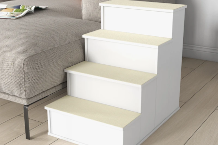 Dog descending white wooden pet stairs beside a large sofa in a bright living room.