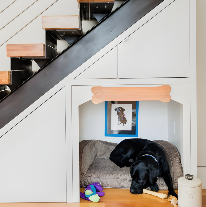 Black dog relaxing in a custom-built bed nook under modern white and wood stairs.