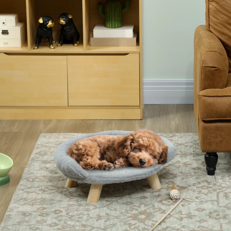 Small dog sleeping on a raised grey round pet bed with wooden legs in a cozy living room.