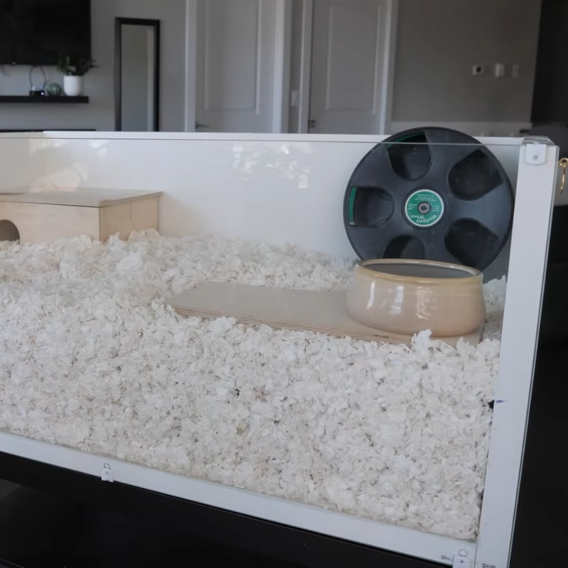 Person pouring sand substrate into a deep-bedding hamster enclosure beside a wooden hideout.