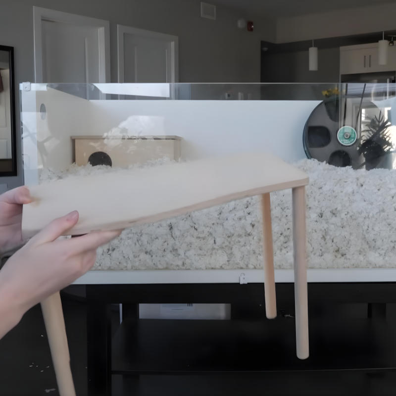 Person holding a wooden platform with legs for stabilizing heavy items in a hamster enclosure.