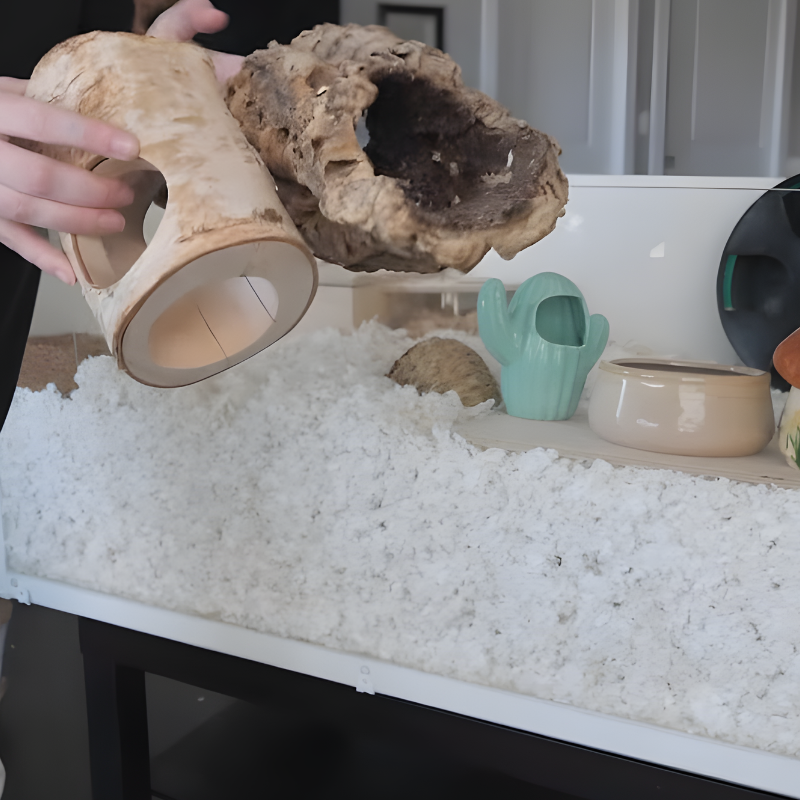 Hand holding natural wooden logs to place inside a deep-bedding hamster cage.