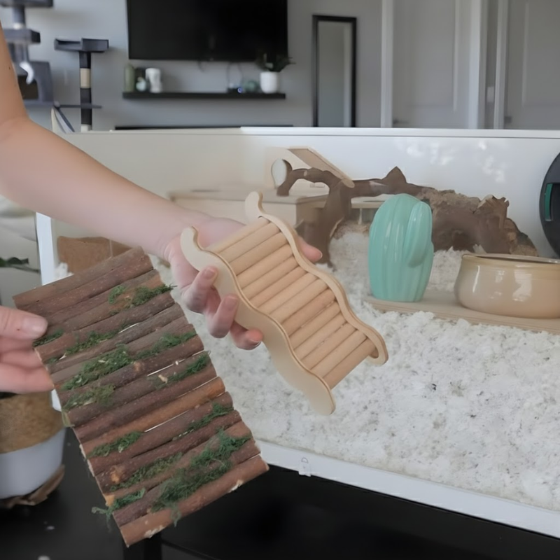 Hand holding two wooden hamster bridges above a deep-bedding enclosure setup.