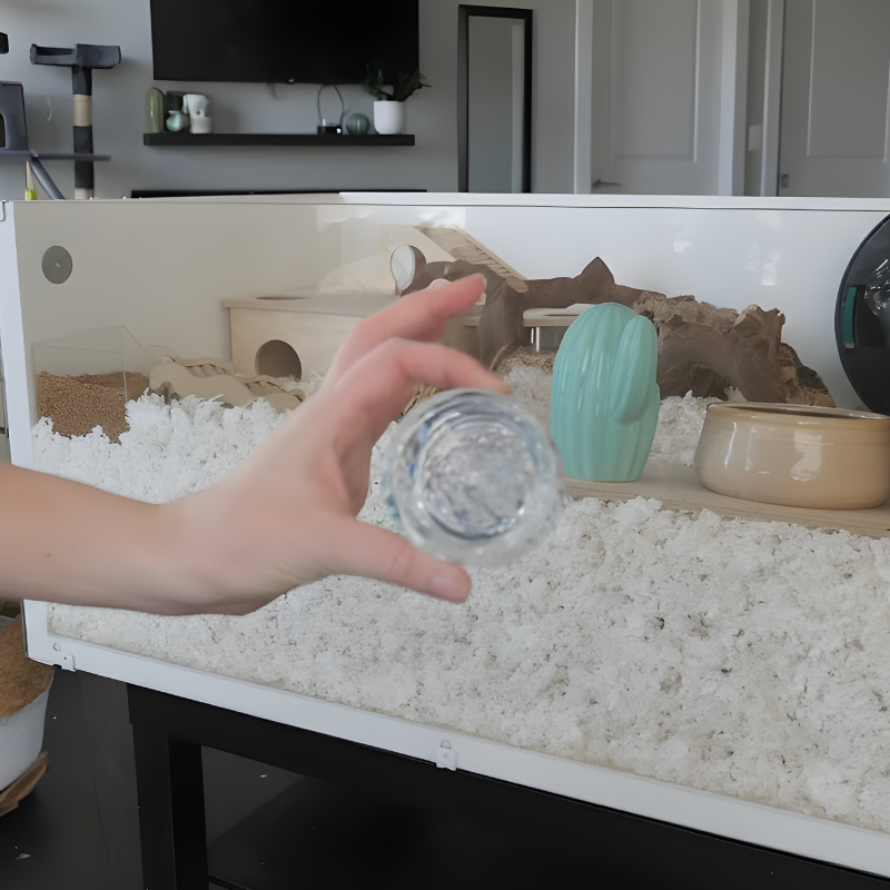 Hand holding a small water container above a deep-bedding hamster enclosure setup.