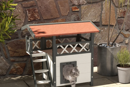 Gray cat exiting multi-level outdoor wooden cat house with potted plants nearby