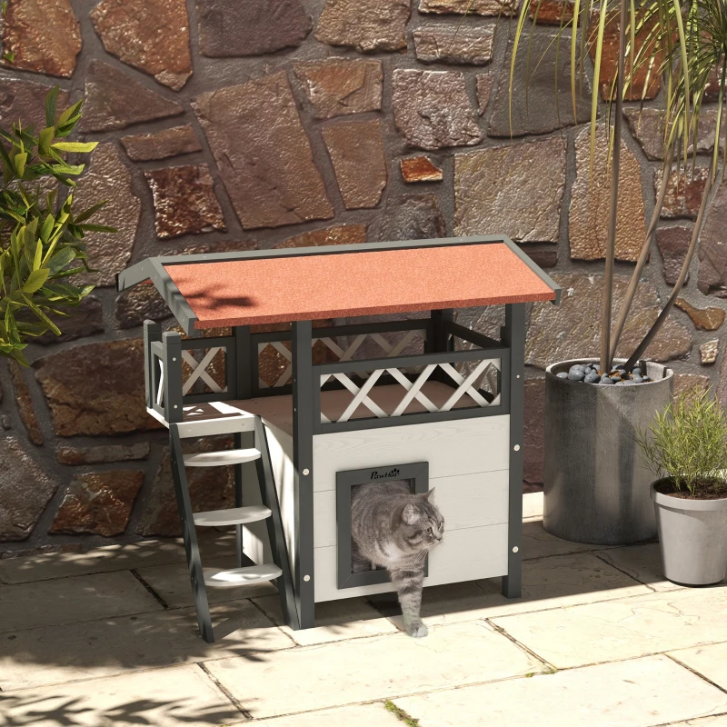 Gray cat exiting multi-level outdoor wooden cat house with potted plants nearby