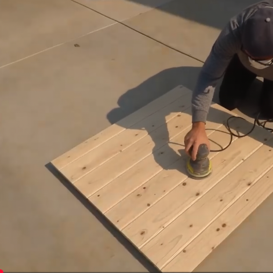 Sanding a flat wooden dog gate panel with an orbital sander for a smooth surface.